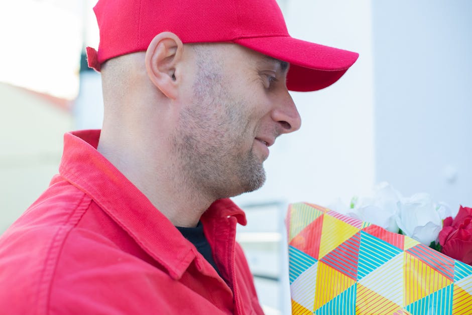 A man wearing a red cap and matching red jacket holds a floral gift wrapped in colorful geometric-patterned paper. The bouquet contains bright pink and white flowers with green stems and leaves, arran