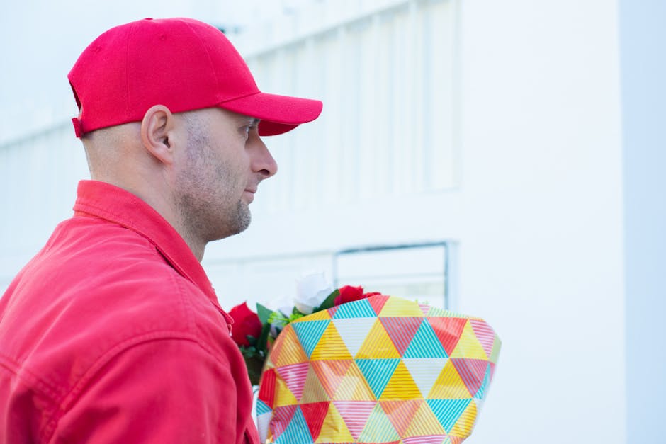 A male flower delivery courier dressed in a red cap and matching red jacket is holding a vibrant floral gift wrapped in geometric-patterned paper featuring yellow, blue, red, and orange triangles. He 