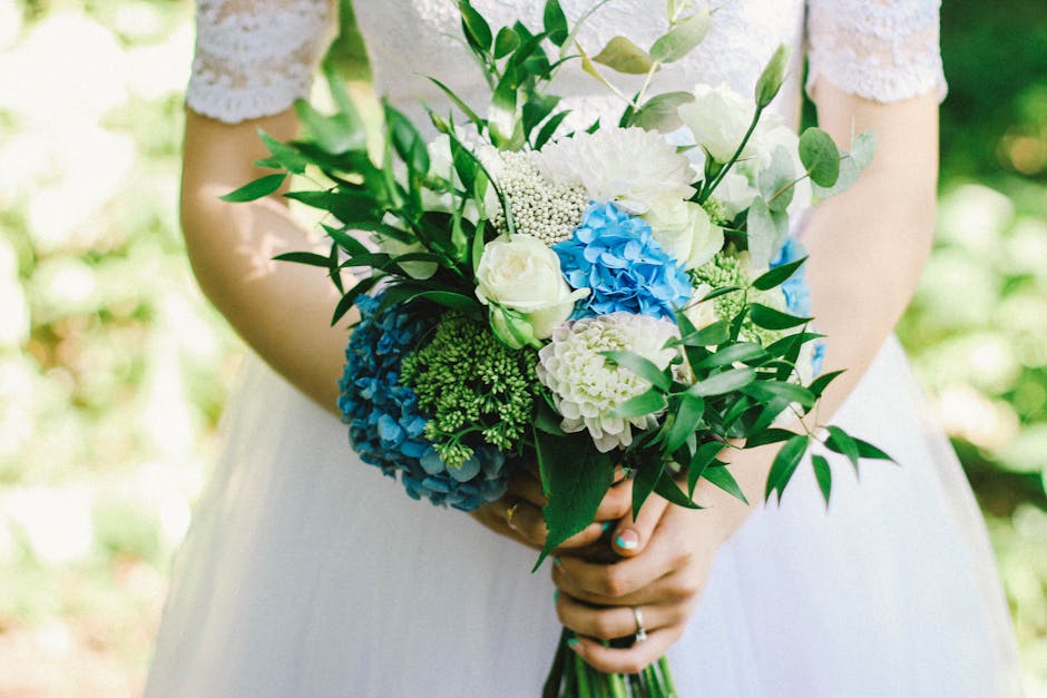 A bride holds a bouquet arrangement featuring white roses, white hydrangeas, blue and green hydrangeas, accented with lush green foliage. The bouquet is wrapped in a natural, light-colored paper and i