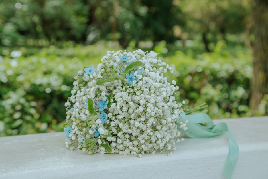 A delicate bouquet of fresh gypsophila flowers, predominantly white with small light blue blossoms interspersed, arranged in a rounded form and tied with a pastel mint-green ribbon. The bouquet rests 