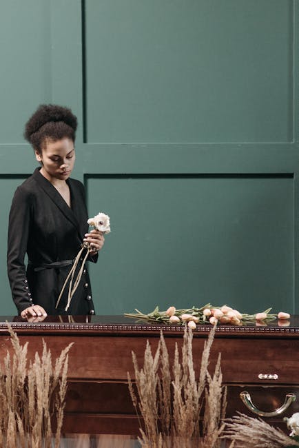 A woman dressed in a black blazer holds a small, delicate white flower floral gift in her hand, standing next to a wooden casket decorated with scattered pale pink roses and green foliage. The setting