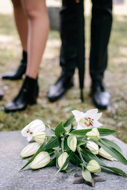 A bouquet of white lilies with large, open blooms and elongated, green leaves sits on a gray stone surface, with closed lily buds in the foreground. In the background, a man and woman, dressed in form