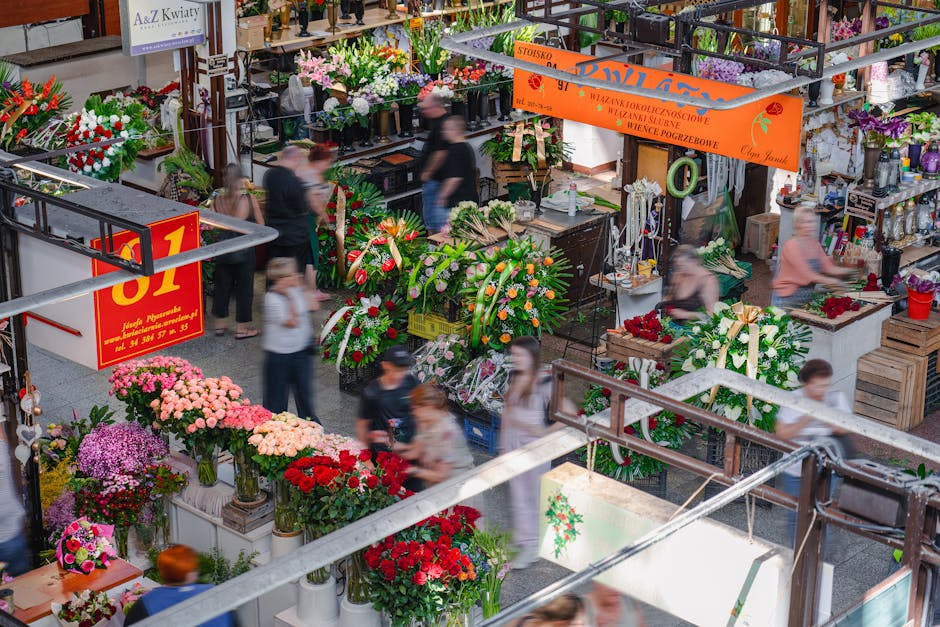 A vibrant flower market scene with a variety of fresh floral arrangements and bouquets displayed across multiple stands. The flowers include pink and purple chrysanthemums, bright red roses, and mixed