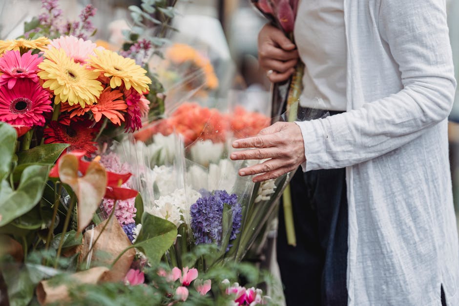 A person wearing a light grey cardigan and dark trousers is selecting a bouquet of fresh flowers from a display at a flower shop on Morden High Street. The floral display includes vibrant pink, yellow