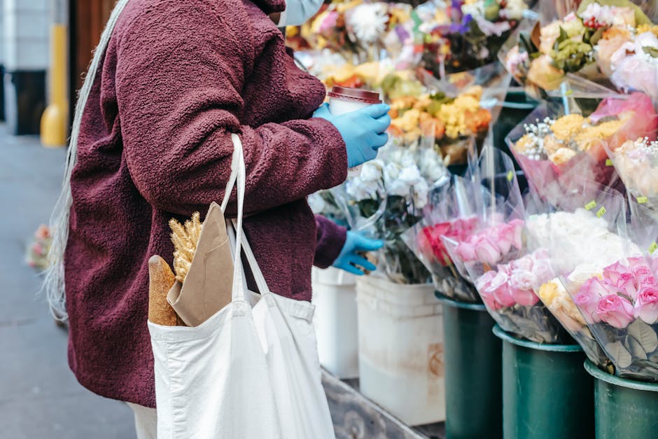 A person wearing a maroon fleece jacket and blue gloves is standing in front of a flower stall on Morden High Street, browsing a variety of fresh floral bouquets. The individual holds a disposable cof